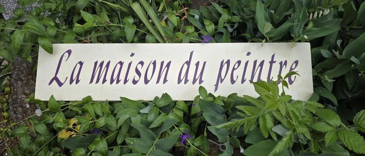 Panneau « La Maison du Peintre » en calligraphie violet aubergine sur fond ivoire, posé dans des feuillages fleuris.