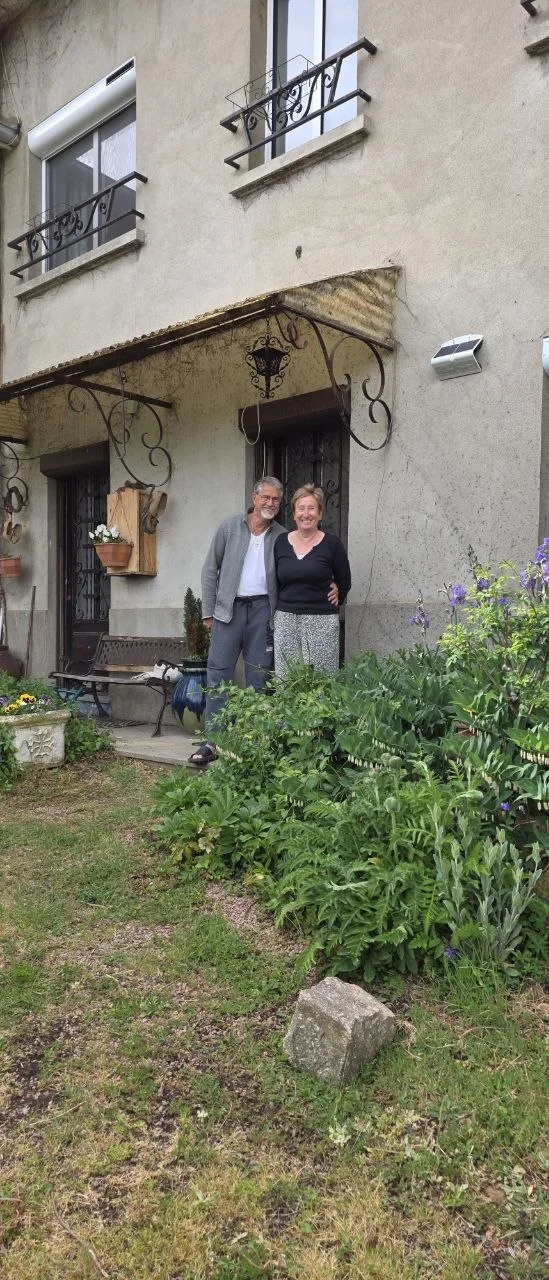 Henri et Marie Odile devant la maison, lui en veste grise et t‑shirt blanc, elle souriant, dans un jardin foisonnant.